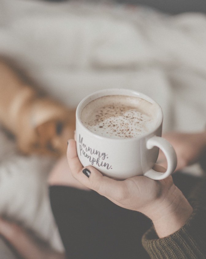 A woman with black nail polish and a green sweater holding a coffee mug of pumpkin spice latte that says "Morning Pumpkin" and