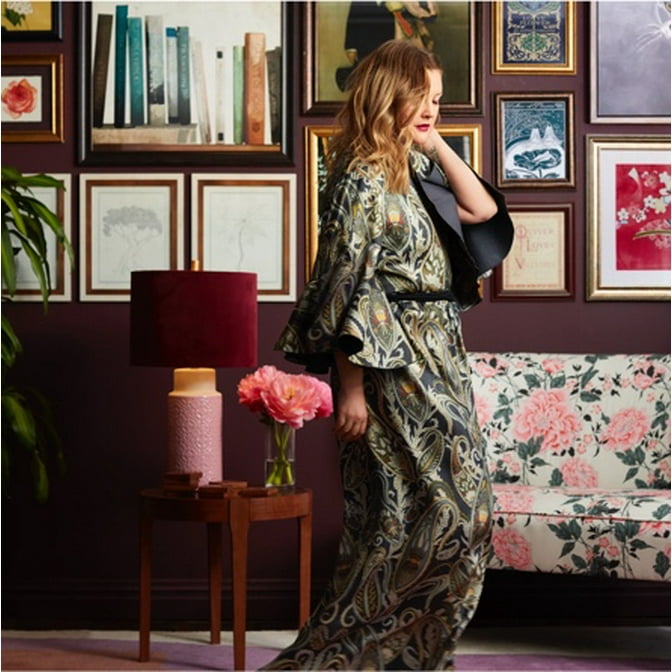 Drew Barrymore in a dark green robe standing in front of a moody modern gallery wall by Drew Barrymore Flower Home and a floral settee.
