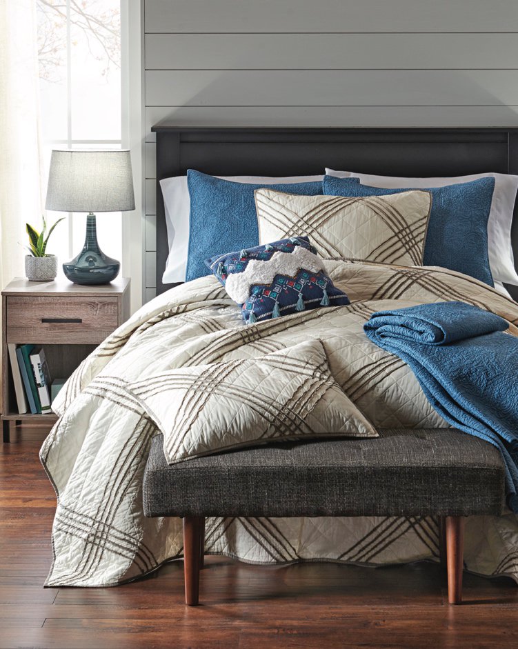 A guest room decorated with Better Homes and Gardens blue and white bedding, a rustic nightstand, a blue table lamp and a chic bench.