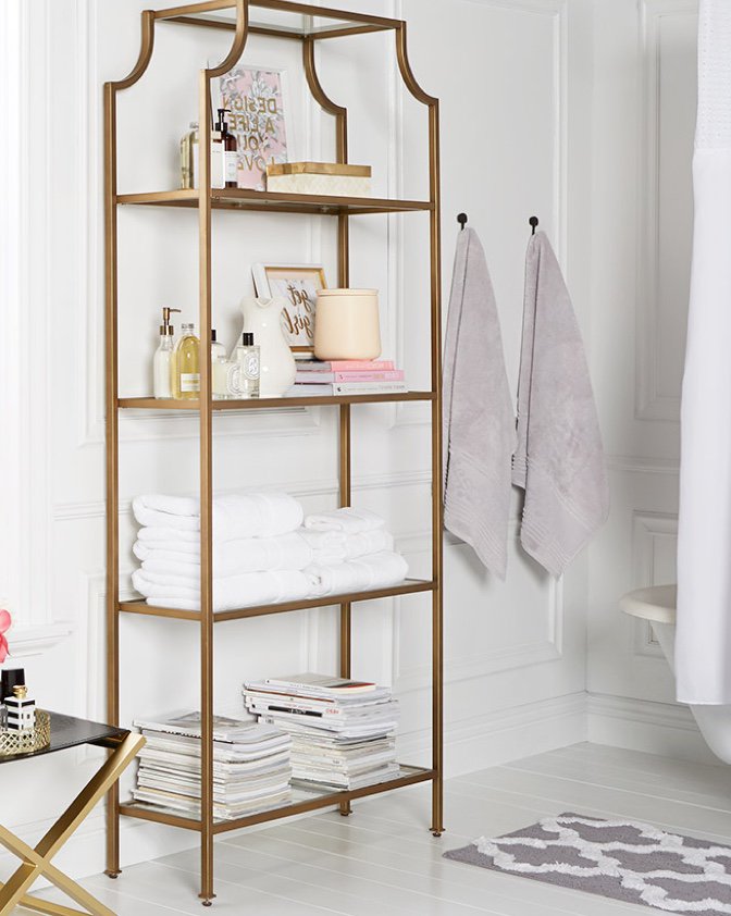 A set of gold shelves in a glam bath with towels hung on the wall and a gold-and-black stool with perfumes displayed in a gold tray.