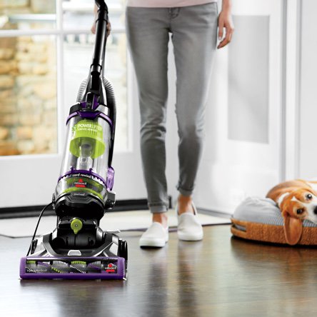 A woman vacuuming with an upright pet vacuum on hardwood floors next to her dog in a dog bed.