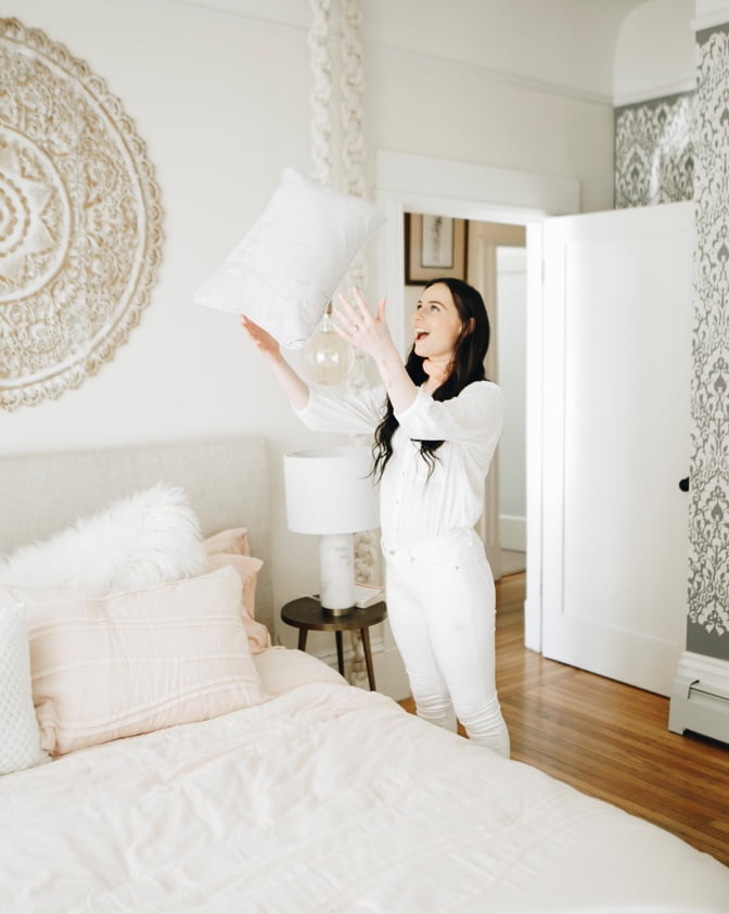 Leigh Winters throwing a MoDRN blush pillow in the air in her airy rustic bedroom.