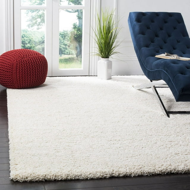 Safavieh Milan Harlow solid white shag area rug displayed in a bright room with dark wood floor, blue tufted lounge chair, red knitted pouf, and potted plant by glass doors.