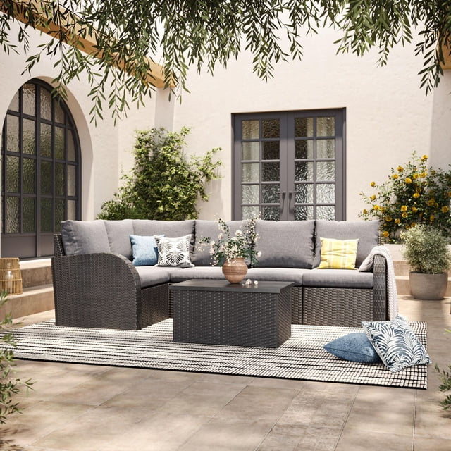 CorLiving Brisbane 6-piece rust-resistant outdoor wicker patio sectional set with gray cushions, matching coffee table, and assorted decorative pillows on a striped outdoor rug in a sunlit courtyard.