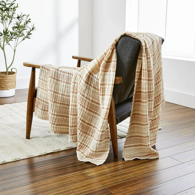 Better Homes & Gardens Striped Gauze Throw, Copper Stripe, 50" x 72", lightweight breathable cotton throw with copper and cream stripes draped over a wooden armchair with dark gray fabric in a bright room with wood floors and a woven basket plant, suitable for year-round comfort.