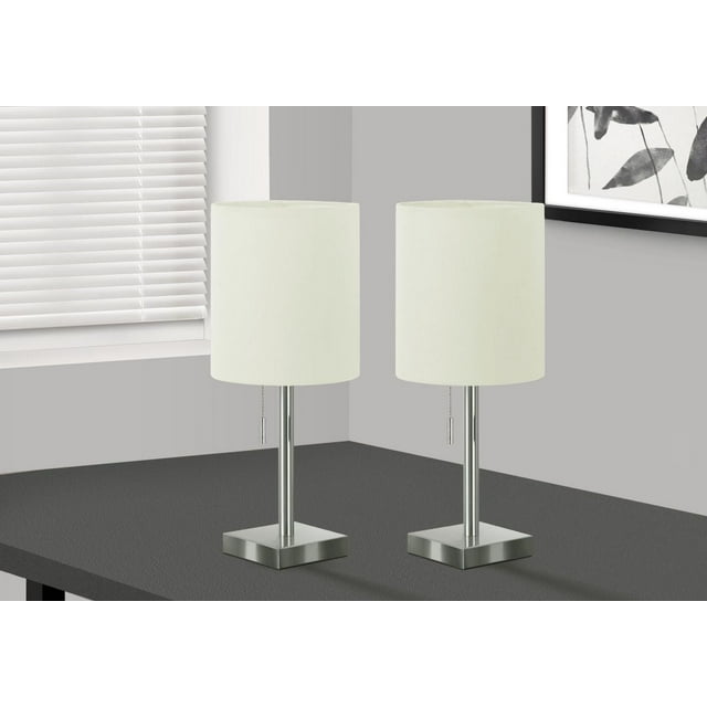 Monarch Specialties Lighting set of 2, 17" contemporary table lamps with nickel metal bases and necks, ivory fabric cylinder drum shades, pull chain switches, and USB ports on the bases, placed on a dark table near a window with white blinds and framed black-and-white wall art, suitable for office or bedroom accent lighting.