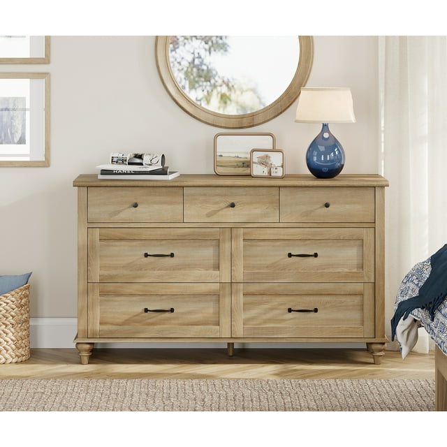 WAMPAT 7-Drawer Dresser in oak finish, featuring a wide wooden chest of drawers with a mid-century modern design. The dresser includes seven spacious drawers with black metal handles, providing ample storage for clothes, blankets, and other items. Crafted from P2 MDF with solid wood legs and an additional middle leg for stability, it is shown in a cozy bedroom setting with a round mirror, a blue lamp, and decorative items on the tabletop. Suitable for bedrooms, kids' rooms, or living rooms.