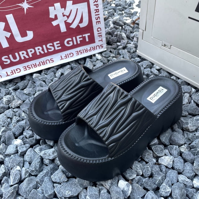zapatillas de mujer con suela gruesa para usar en verano, exquisitas sandalias de moda, zapatos de playa: par 3/4 frontal de sandalias negras de plataforma con suela gruesa y plantilla EVA, banda superior texturizada con pliegues, tacón alto aprox. 9 cm y punta abierta; colocadas sobre grava junto a una caja con texto.