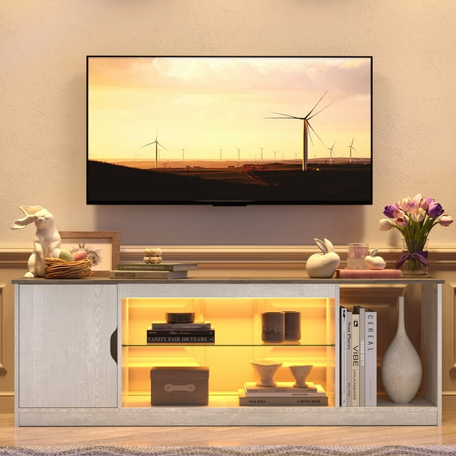 Bestier 58" LED TV Stand for PS5 Gaming Entertainment Center for 60/65 inch TV in wash white finish. This modern TV console features a closed cabinet on the left, adjustable tempered glass shelves with RGB LED lighting in the center, and open storage on the right. Designed for TVs up to 65 inches, it provides ample space for gaming consoles, controllers, and accessories. Displayed in a cozy living room setting with decorative items and a mounted TV above.