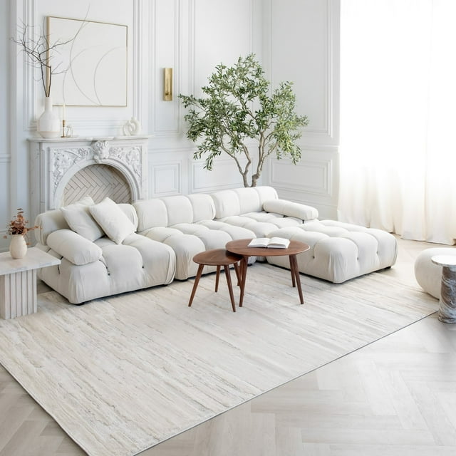 Modern beige cream abstract indoor area rug placed under a white tufted sectional sofa and wooden coffee tables in an elegant, bright living room setting.