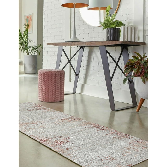 Amber traditional runner rug featuring intricate beige and rust patterns on polished concrete floor under a wooden table in a bright living space with white brick walls, green plants, round mirror, lamp, and pink knitted pouf.