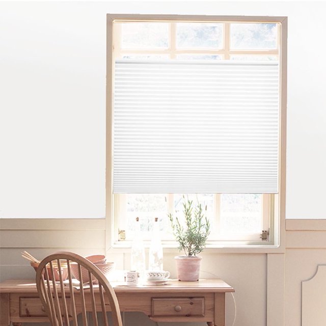 persianas plisadas autoadhesivas de 90 x 150 cm, semiopacas, para baño, ventanas, cortinas, textiles para el hogar, color blanco — vista frontal centrada de cortina plisada blanca 90 x 150 cm instalada en ventana; pliegues horizontales uniformes y acabado semiopaco, borde superior con cinta autoadhesiva visible, caída parcial sobre alféizar con escritorio de madera, maceta y silla; adecuada para controlar luz y privacidad en baños o ventanas pequeñas y opción frecuente para instalación sin taladro.