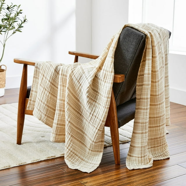 Better Homes & Gardens Striped Gauze Throw beige stripe, 50" x 72", lightweight breathable cotton throw draped over a wooden armchair with dark gray cushions in a bright room with hardwood floors, cream rug, and a potted plant, ideal for year-round comfort.