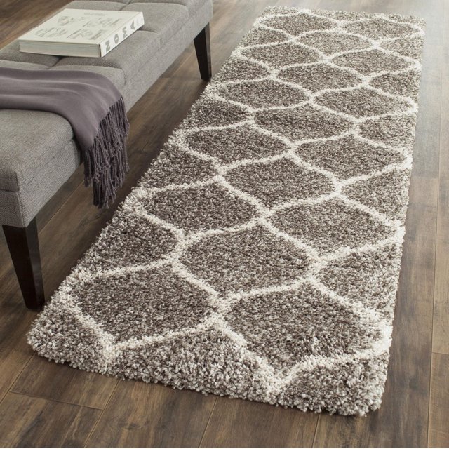 Safavieh Hudson Arline geometric shag area rug in beige and cream tones placed on wood flooring beside a gray upholstered bench with a book and purple throw.