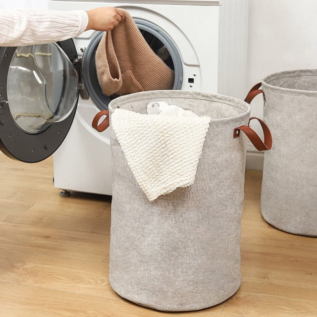 cesta grande cilíndrica gris para ropa sucia, cesta de almacenamiento de tela teñida con hilo, cesta plegable impermeable para ropa, cesta de almacenamiento de tela con doble asa — Vista 3/4 frontal de una cesta cilíndrica gris (50 x 36 cm) en cuarto de lavado; tela teñida con textura visible, borde superior rematado y asas dobles marrón reforzadas, con una toalla clara sobresaliendo; lavadora y otra cesta al fondo. A menudo usada también para guardar artículos sueltos en baño o cuarto de lavado.