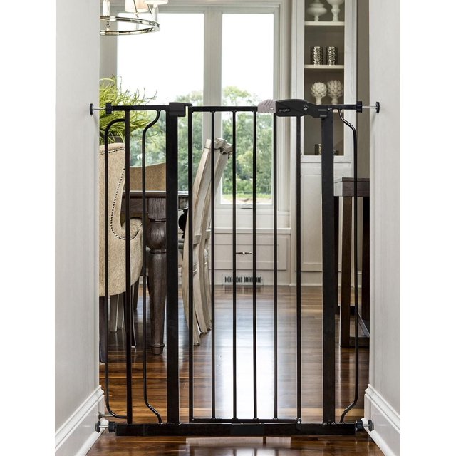 Regalo Extra Tall Baby Gate Black, a sturdy all-metal safety gate with vertical bars and pressure mount assembly, fits openings 29 to 36.5 inches wide and stands 41 inches tall. It features a walk-through door with an easy lever handle and one-touch release safety lock, installed in a hallway with hardwood floors and dining furniture visible behind.