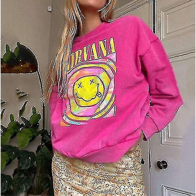 nirvana smiley face crewneck sudadera heliconia color nirvana sudadera regalo sudadera rosa heliconia talla XXL de spandex con cuello redondo y estampado frontal amarillo de cara sonriente y texto NIRVANA; vista 3/4 frontal, manga larga y puños acanalados sobre falda en interior con plantas. A menudo usada como suéter informal para conjuntos de moda urbana.