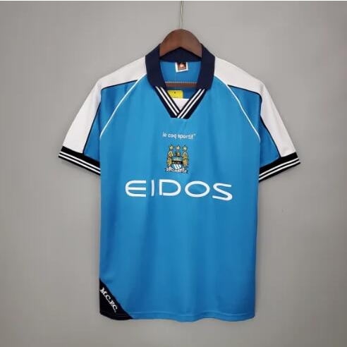 jersey retro 99-01 manchester city home camiseta retro Manchester City 1999-2001 azul celeste con mangas blancas y ribetes negros, cuello en V azul marino con doble raya blanca y negra, escudo del club y logo EIDOS impresos en el pecho; vista frontal colgada sobre percha con fondo neutro. talla XL mostrada; suele usarse como pieza de colección o para uso casual.