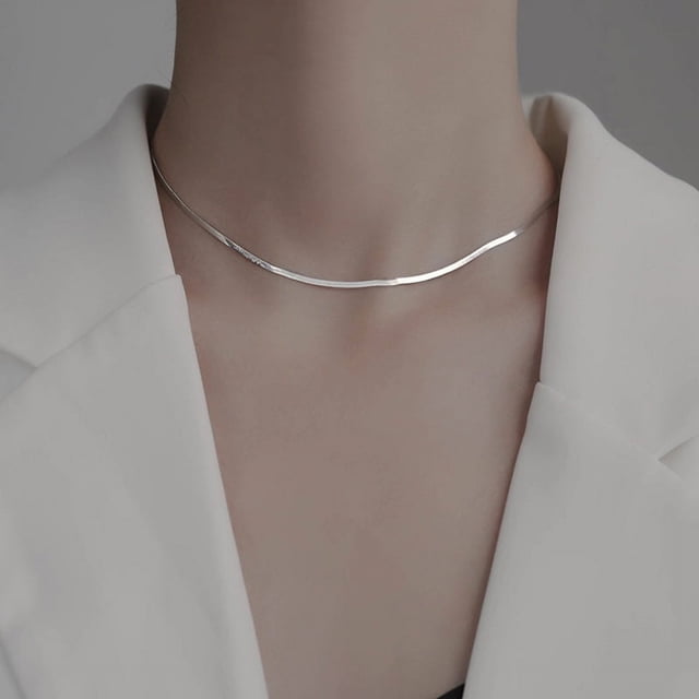 cadena de hueso de serpiente plana de plata s925, collar de cuchillo estilo frío; gargantilla femenina plateada visible sobre la clavícula en vista frontal, cadena plana y continua con acabado brillante y eslabones lisos, modelo con saco claro en fondo neutro, a menudo usada sola o en capas.