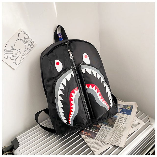 bape shark mochila escolar trend personalizada graffiti estudiante bolsa de deporte negra 26 pulgadas, vista frontal centrada con cierre vertical que forma boca de tiburón; dientes blancos serrados e interior rojo, ojillos blancos con puntos rojos en la parte superior, asa y correas negras, acabado brillante, apoyada sobre maleta de aluminio en un rincón minimalista; a menudo usada como mochila escolar o para llevar equipo deportivo ligero.