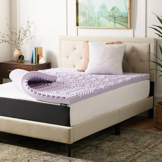 lucid - colchoneta de memory foam con infusión de lavanda - sobrecolchón de 5 zonas de 7.5 cm - topper para colchón - sensación suave y acolchada - alivia puntos de presión - matrimonial vista 3/4 del sobrecolchón matrimonial lila de 7.5 cm parcialmente enrollado sobre un colchón; superficie texturizada con canales de ventilación y relieves que definen las 5 zonas de soporte para cabeza, hombros, espalda, caderas y piernas; tejido exterior liso y color lavanda claro; dormitorio con cabecera y mesita de noche al fondo. Se coloca directamente sobre el colchón y se usa con protector y sábana ajustable; también suele utilizarse para añadir confort extra en colchones firmes durante viajes.