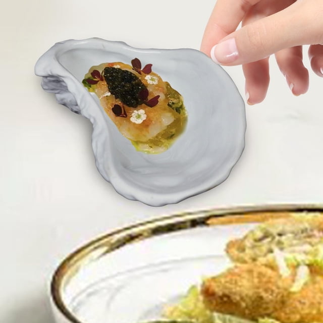 plato de cerámica para ostras, plato con forma de concha de ostra, creativo, apto para lavavajillas, plato de aperitivo de duradero para plato de cerámica blanca en vista 3/4, 13.5 x 7 x 4.2 cm, contorno ondulado que imita una valva y superficie lisa; mano sosteniendo una porción de ostra con aderezo. Ideal para servir ostras y pequeños aperitivos; también suele usarse para presentar bocados en menús de restaurante.