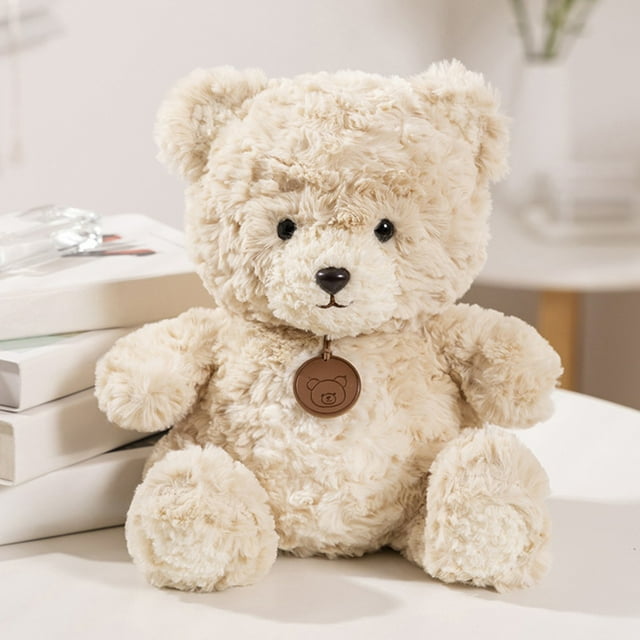 TIMIFIS Plush Doll 13.8 Inch Teddy Bear, a classic teddy bear plush toy made of soft polyester material, shown in a beige color with a textured short plush exterior. The bear features small black eyes, a dark brown nose, and a round medallion with a bear design hanging from its neck. Positioned on a white surface with books in the background, this stuffed animal is ideal as a soothing children’s doll, holiday or birthday gift, or decorative item for a dorm, RV, or home office.