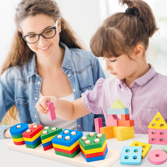 figuras geométricas de madera para bebés juego de clasificación de matemáticas Vista frontal; niño pequeño manipulando piezas geométricas apilables de madera en colores rojo, azul, verde, amarillo y rosa sobre base de tablero; incluye cuadrados, círculos, triángulos y bloques con clavijas; acabado lijado, esquinas redondeadas y pintura a base de agua; apto para 1 año+.