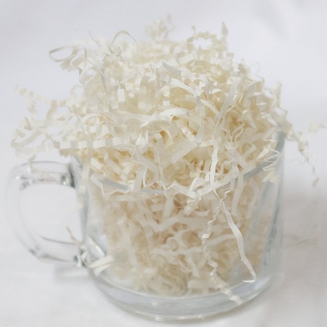papel triturado blanco de 500 g, engarzado suave para embalaje: tiras de papel de seda blanco crudo en una taza de vidrio, vista frontal; textura fina y arrugada, corte en tiras rizadas y aspecto mate, usado como relleno decorativo para cajas, cestas y regalos; a menudo usado en manualidades y presentaciones festivas.