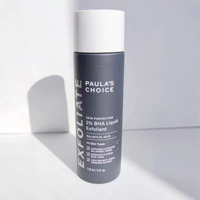 paula's choice skin perfecting - líquido salicílico exfoliante 2% bha, botella cilíndrica gris con tapa blanca en vista frontal, texto del envase que indica 2% BHA y ácido salicílico; superficie mate con sombra lateral sobre fondo blanco minimalista; indicado para puntos negros y poros grandes, apto para todo tipo de piel; también suele usarse para preparar la piel antes de tratamientos faciales.