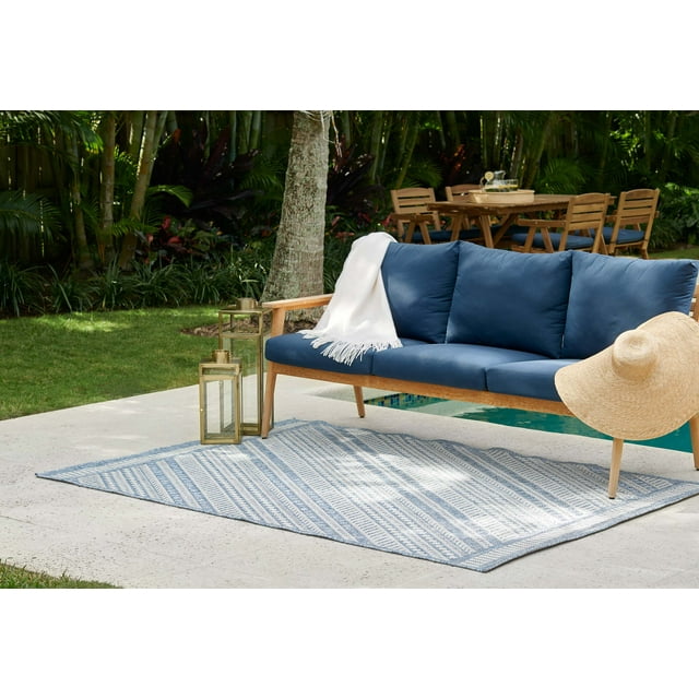 transitional outdoor area rug under blue cushioned wooden sofa with white throw and straw hat beside brass lanterns near pool