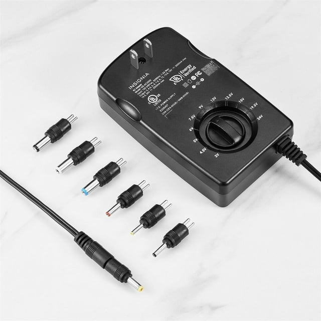 Insignia 3,000 mA Universal AC Adapter for High-Output Devices 3 to 24 Volt, shown with a black rectangular body featuring a manual voltage selector knob and LED voltage indicator. Includes seven interchangeable round-pin connector tips in various sizes and colours, suitable for devices requiring 3–24 volts. The adapter has a 5 ft. attached cord for flexible placement and is designed for high-output devices up to 3,000 mA. Positioned on a white surface.