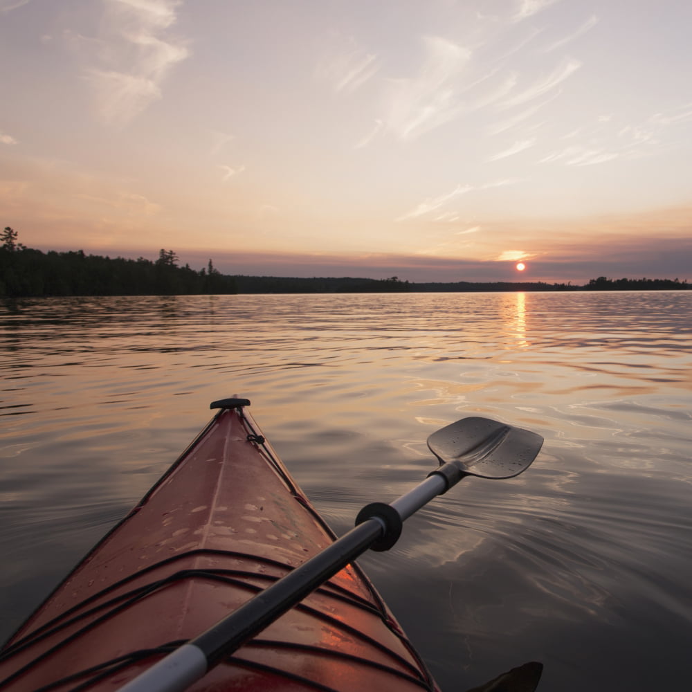 Bow of a red kayak and paddle on a tranquil lake at sunset;Keewatin ...