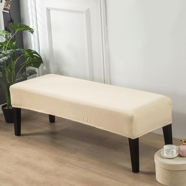 cubierta de banco de poliéster tejido jacquard beige macarena funda de asiento de banco elástica, vista frontal centrada de un banco rectangular con funda beige texturizada, tejido jacquard y dobladillo elástico cosido visibles que ajustan la base; se ven patas de madera oscura y fondo de sala con planta y pared blanca. Adecuada para bancos de 107–137 cm de largo y 36–46 cm de ancho.