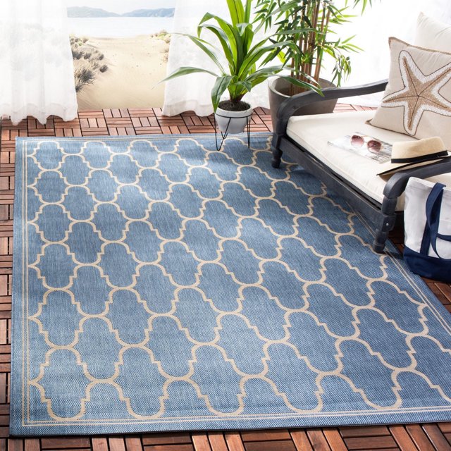 SAFAVIEH Courtyard Eustacia blue geometric indoor/outdoor area rug on wood tile patio floor next to white cushioned bench with starfish pillow and green plants.