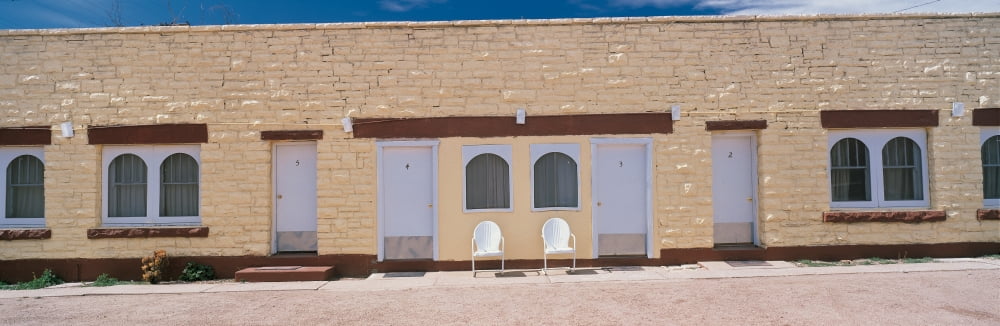 This is a yellow brick motel on Route 66 it sits next to the road There are two chairs in front of the motel There are windows spaced across the motel and closed doors to the hotel rooms Canvas Art -