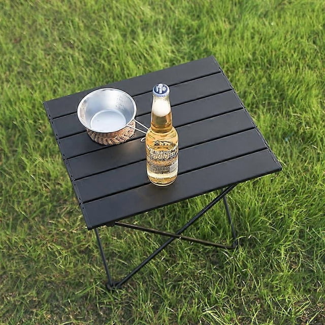 mesa plegable ligera de aleación de aluminio para acampar, picnic, mesa pequeña para exteriores, equipo para exteriores. Mesa cuadrada plegable con tablero de lamas negras y estructura de aluminio en X, vista 3/4 desde arriba; botella y cazo sobre la superficie, patas con base antideslizante y, según especificaciones, bolsillo de almacenaje en tela Oxford; tamaño pequeño 41x35x30cm.