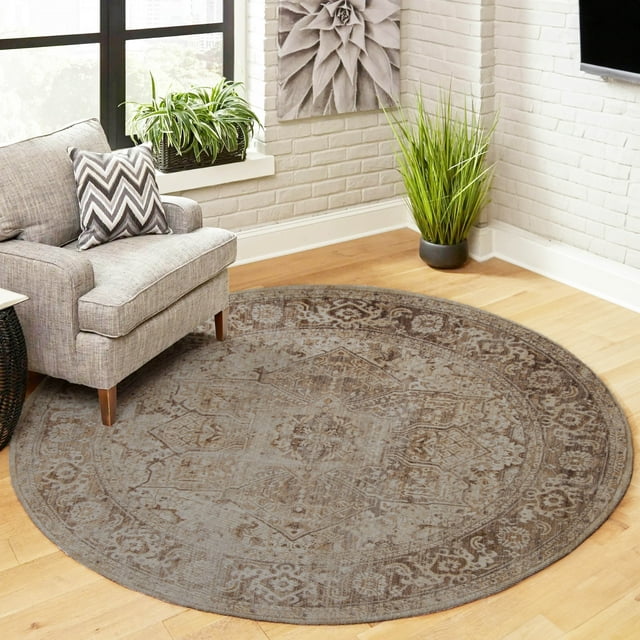 Round ECARPET Selena traditional area rug with muted beige and brown patterns on hardwood floor beside a gray upholstered armchair, near white brick walls and indoor plants.