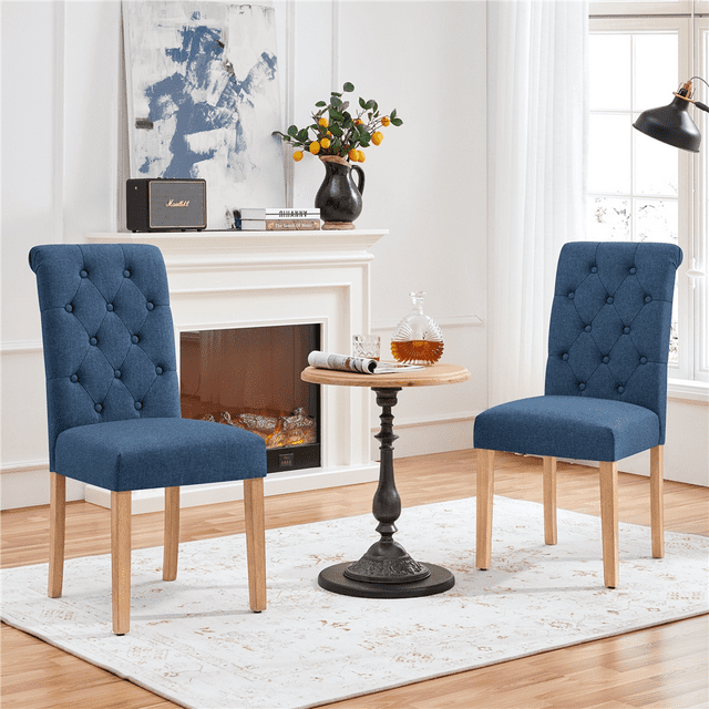 Yaheetech Upholstered Padded Dining Chairs with Adjustable Foot Pads, Set of 2, Blue. These dining seating options feature button-tufted linen-like fabric, curved rear legs, and durable rubberwood construction. The high-density foam seat cushions ensure comfort, while adjustable foot pads provide stability on uneven floors. Ideal for kitchen or dining spaces, shown in a cozy room with a fireplace and a small round table.