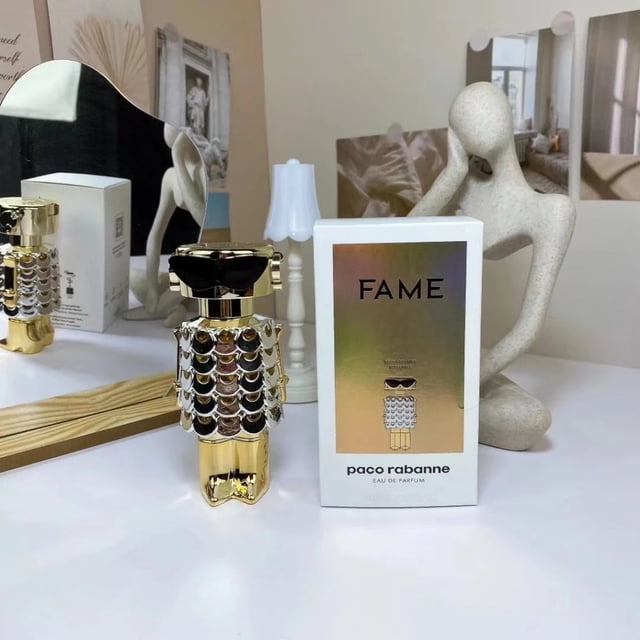 contador paco rabanne 2022 nuevo producto robot señoras perfume fuerte fama edp 80ml botella dorada con detalles tipo escamas y forma estilizada de robot, caja blanca con portada iridiscente "FAME" y texto paco rabanne, vista frontal en ángulo sobre mesa con espejo y figura decorativa de fondo; eau de parfum para mujer, presentación 80 ml.