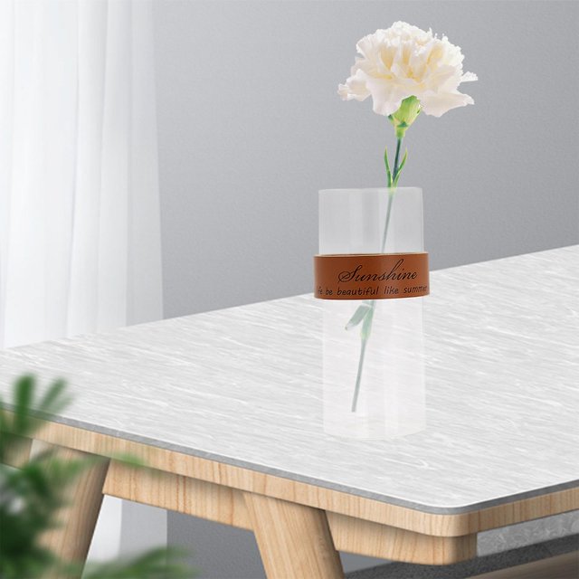 florero de vidrio transparente sharpla con correa de cuero marrón — Centrado, vista frontal de un jarrón cilíndrico de vidrio transparente con correa de cuero marrón con texto alrededor del cuerpo; contiene un clavel blanco seco, borde soplado a mano visible, tamaño aproximado 8x18 cm, sobre mesa clara en ambiente interior minimalista; adecuado para flores secas y decoración de sala o cafetería.
