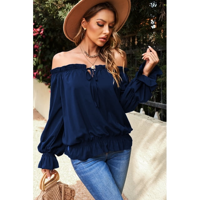 abphqto blusa azul marino con hombros descubiertos y manga abullonada con volantes para mujer blusa azul marino, 3/4 frontal sobre modelo; escote off-shoulder elástico con lazo delantero, mangas largas abullonadas con volantes en los puños, bastilla elástica fruncida y caída fluida, combinada con jeans en ambiente exterior.