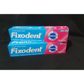 fixodent original 68 g (kit con 02 unidades) kit de dos tubos de crema fijadora para prótesis dentales, vista frontal apilada en empaque azul y rosa con logotipo Fixodent y texto 'Original', cada tubo 68 g; según especificaciones reduce molestias por roce de encías y no interfiere con el sabor; adecuado también para llevar en viajes, fondo negro.