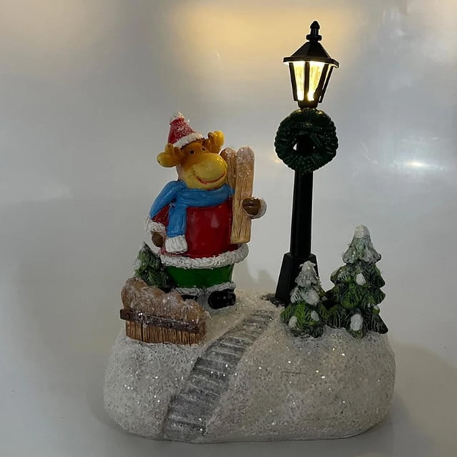 decoración de mesa navideña macarena iluminada figura navideña led lámpara de calle brillante, vista frontal centrada de una microescena sobre base nevada con farola LED encendida, alce con gorro y bufanda sosteniendo esquís, dos pinos con nieve y escalones tallados; resina pintada, detalle fino, tamaño compacto ideal como centro de mesa o adorno de repisa, a menudo usado también como adorno de escritorio festivo.
