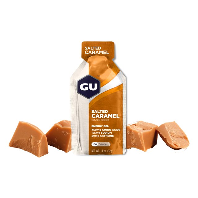 gu energy labs original, gel energético de nutrición deportiva, caramelo salado, caja de 24 piezas, vista frontal centrada de un sobre monodosis color caramelo y blanco con logo GU; texto legible 'Salted Caramel', 'Energy Gel', '100 calories', 'aminoácidos de cadena ramificada', '125 mg sodium', '20 mg caffeine', sobre fondo blanco con trozos de caramelo salado; indicado para entrenamiento o carreras y a menudo usado como gel portátil durante la actividad física.