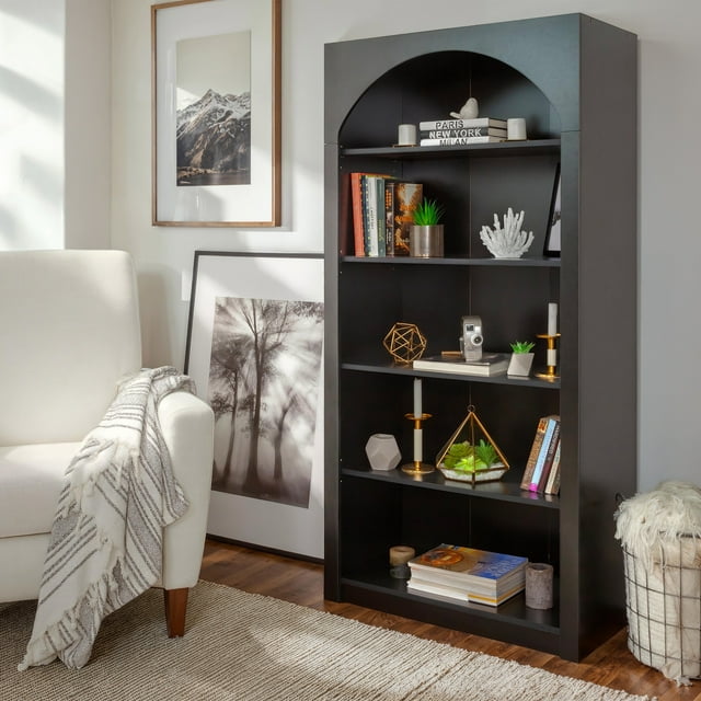 Black CorLiving Caldwell engineered wood bookcase with arched top featuring five shelves holding books, plants in pots and terrariums, candles on gold holders, vintage camera decor piece; placed beside white armchair draped with striped throw blanket; framed black-and-white nature photos leaning against wall on wooden floor.
