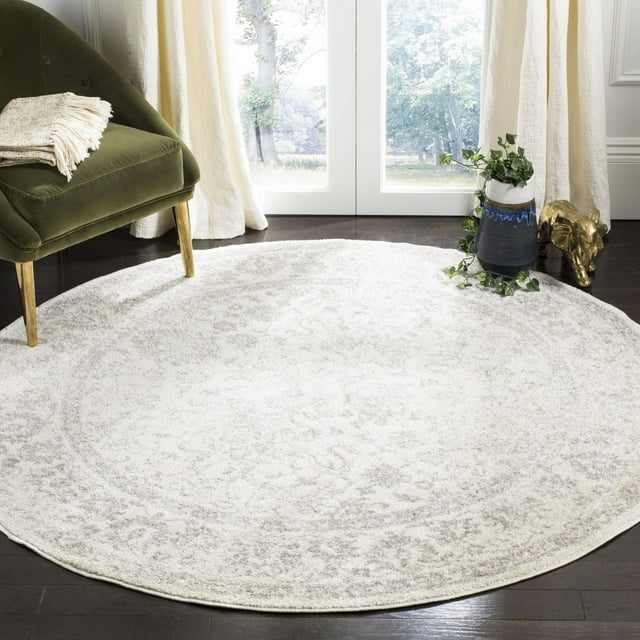 SAFAVIEH Adirondack Wyatt round traditional area rug with subtle floral design in light gray and cream, placed on dark hardwood floor near green chair and decorative vase.