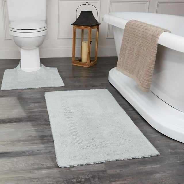 Set of two light gray Fabstyles Basix soft and absorbent reversible cotton bath rugs displayed on dark wood floor near white toilet and bathtub with beige towel.