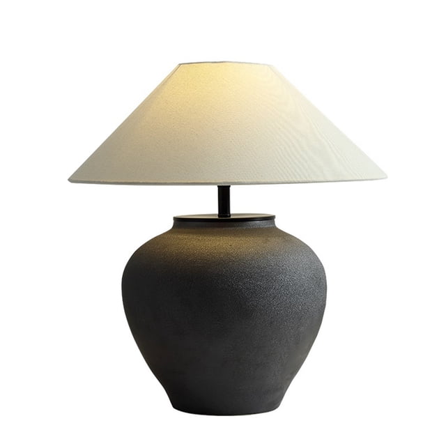 lámpara de mesita de noche de cerámica hecha a mano, base negra texturizada tipo maceta y pantalla de lino crema; vista frontal centrada, silueta redondeada, difusión de luz suave para crear una atmósfera cálida; a menudo se usa también como adorno decorativo en estantes o consolas.