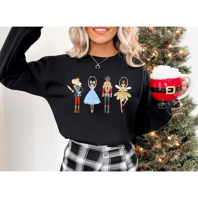 sudadera navideña de cascanueces para mujer, jersey de hada de ciruela de azúcar, camisa de escuadrón de cascanueces, ballet, ropa linda de invierno Negra sudadera centrada, vista frontal con cuatro figuras estampadas de cascanueces y bailarinas en el pecho; cuello redondo, mangas largas y corte relajado, modelo frente a un árbol de Navidad iluminado sosteniendo una taza temática; a menudo usada como prenda festiva para eventos informales.